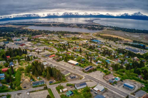 Aerial view of a small town surrounded by greenery and mountains, with a lake and cloudy sky in the background.