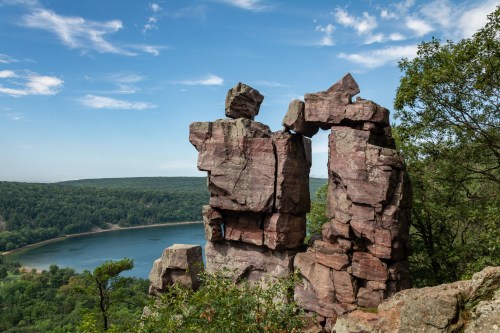 Rock formations stand tall against a blue sky, overlooking a serene lake surrounded by lush greenery.