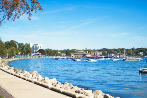 A scenic lakeside view with boats docked, a clear blue sky, and a pathway lined with rocks and trees.