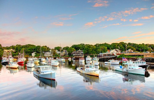 A serene harbor at sunset, featuring various boats moored in calm waters, surrounded by lush greenery and quaint buildings.