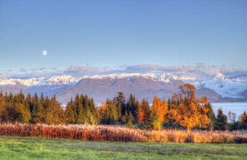 A serene landscape featuring snow-capped mountains, autumn trees, and a full moon in a clear blue sky.