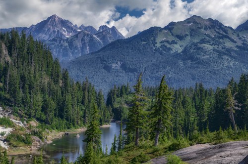 A serene landscape featuring a river surrounded by lush green trees and majestic mountains under a partly cloudy sky.