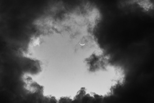 A crescent moon peeks through a circular opening in dark, cloudy skies.