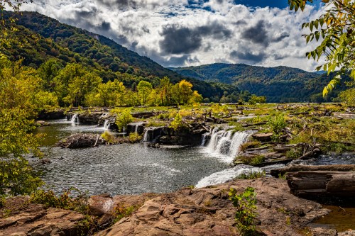 A serene landscape featuring a waterfall cascading into a calm river, surrounded by lush greenery and mountains under a cloudy sky.