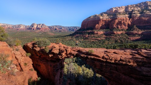 A person stands triumphantly on a rock arch, overlooking a vast red rock landscape in Sedona, Arizona.