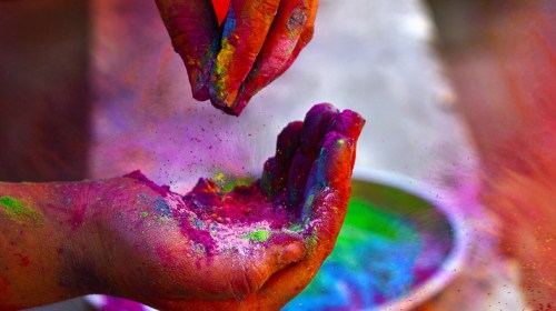 A close-up of hands covered in vibrant colored powders, sprinkling them over a bowl filled with more colors.