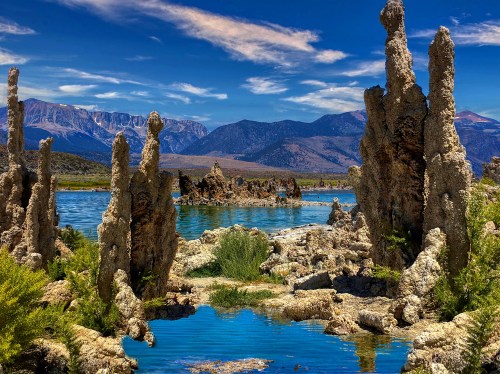 Scenic landscape featuring unique rock formations by a blue lake, surrounded by mountains and a clear blue sky.