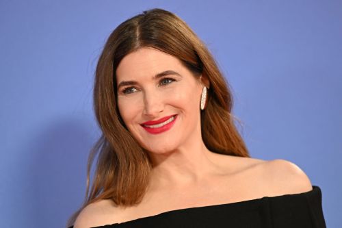 A woman with long brown hair smiles, wearing an off-the-shoulder black top and bold red lipstick against a blue background.