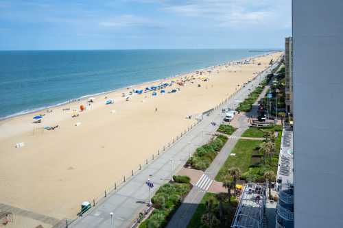 A wide view of a sandy beach with colorful umbrellas, a boardwalk, and calm blue ocean under a clear sky.