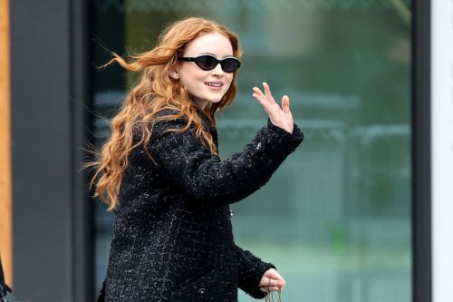 A smiling person with long, wavy red hair waves while wearing sunglasses and a black coat.