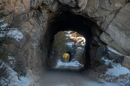 A dirt road passes through rocky tunnels, with sunlight illuminating the distant exit in a snowy landscape.