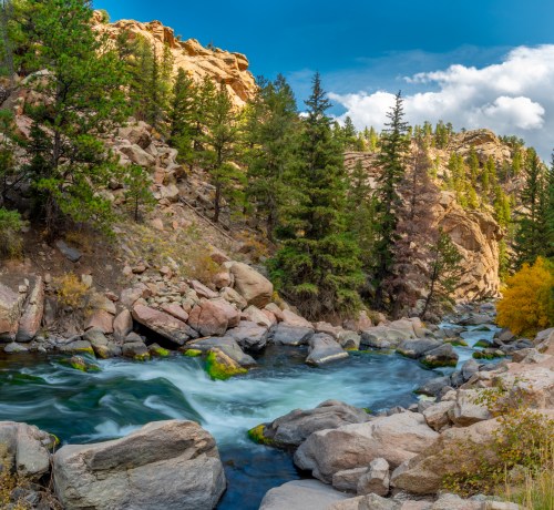 A serene river flows through rocky terrain, surrounded by lush green trees and golden foliage under a blue sky.