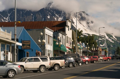 A scenic view of a small town street lined with parked cars, surrounded by mountains and cloudy skies.