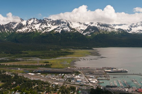 A scenic view of snow-capped mountains overlooking a coastal town and marina, with lush greenery and a cloudy sky.