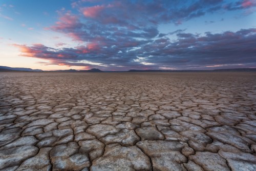 A vast, cracked dry landscape under a colorful sky at sunset, with distant mountains on the horizon.