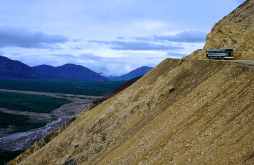 A bus travels along a steep, rocky road with mountains and a river in the background under a cloudy sky.