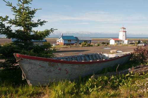 A weathered boat in the foreground with a lighthouse and buildings in the background, set against a scenic landscape.