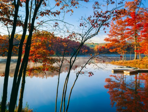 A serene lake surrounded by vibrant autumn foliage and mist, with a small dock reflecting in the calm water.