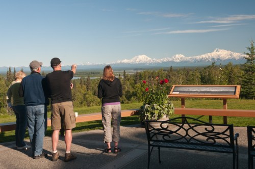 A group of people stands on a viewpoint, admiring a scenic mountain landscape with flowers and a bench nearby.