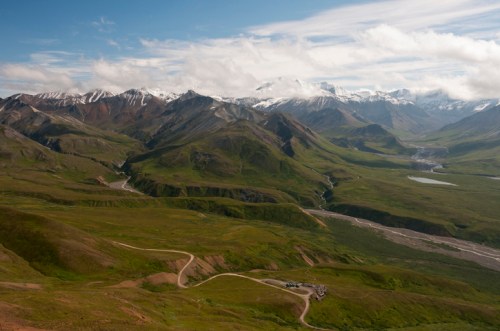 A panoramic view of lush green mountains with snow-capped peaks under a blue sky and scattered clouds.