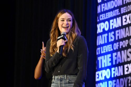 A smiling woman holds a microphone on stage, with a backdrop featuring event branding and text about fandom.