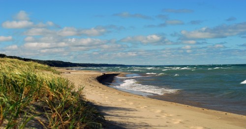 A sandy beach with gentle waves, grassy dunes, and a blue sky filled with clouds.