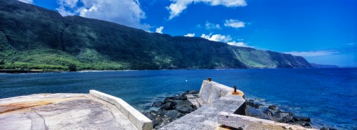 A scenic view of a rocky coastline with lush green mountains under a bright blue sky and calm ocean waters.