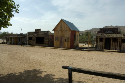 A deserted old western town with wooden buildings, including a jail, set against a dusty landscape and mountains.