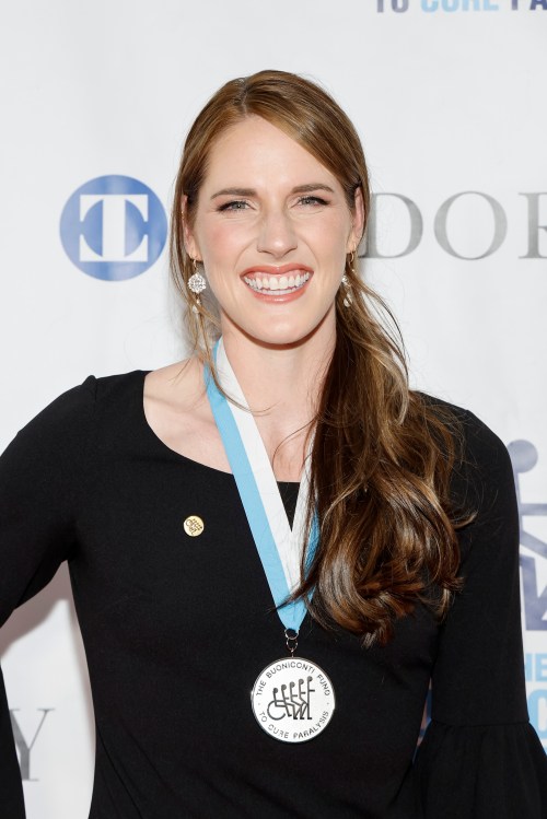 A smiling woman in a black dress wearing a medal, posing at an event with a backdrop featuring logos.