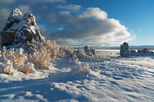 Snow-covered landscape with rocky formations and sparse vegetation under a bright blue sky and fluffy clouds.