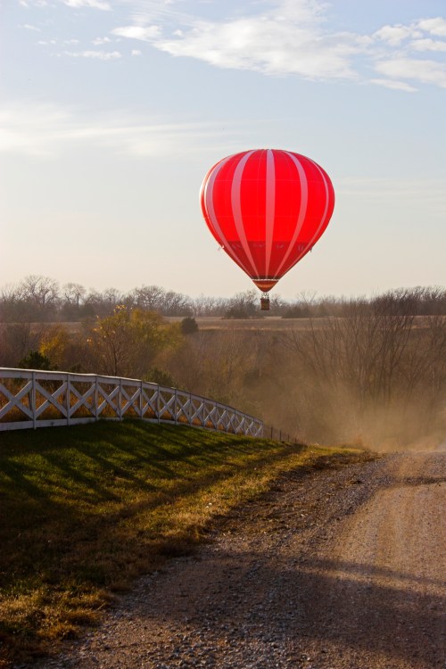 A red hot air balloon floats above a gravel path, surrounded by trees and a clear sky.