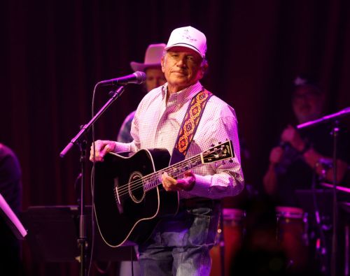 A musician in a white cap plays an acoustic guitar on stage, singing into a microphone with a band in the background.