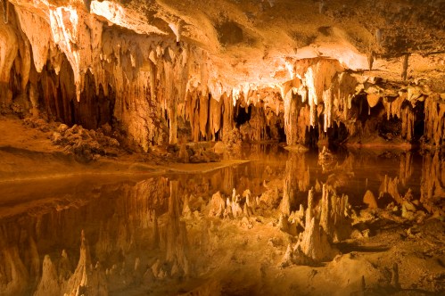 A stunning cave interior with stalactites and stalagmites reflected in still water, illuminated by warm lighting.