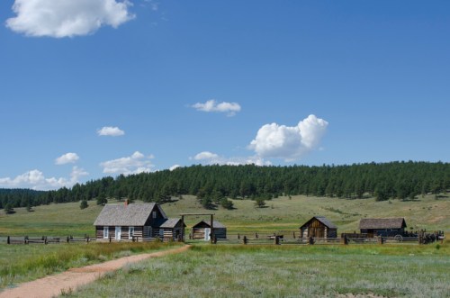 A rustic homestead with log cabins set against a green landscape and blue sky, surrounded by trees and open fields.