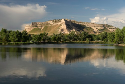 A serene lake reflects a rocky cliff and lush greenery under a blue sky with scattered clouds.