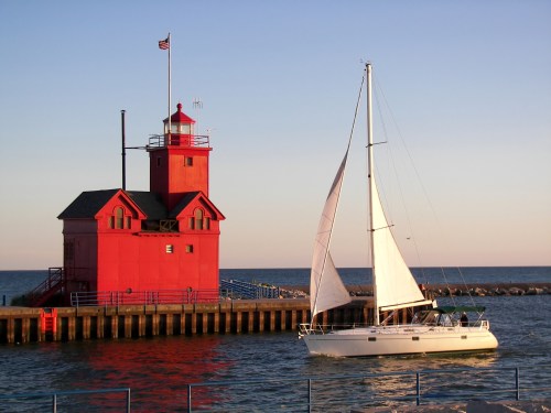 A red lighthouse stands by the water as a sailboat glides past during sunset.