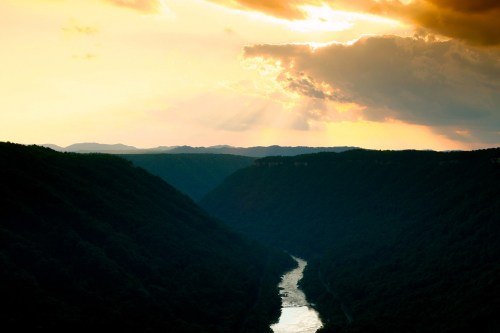 A serene river flows through a valley, framed by mountains under a dramatic sunset sky with rays of light.