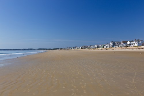 A sandy beach stretches along the shore, with houses lining the coast under a clear blue sky.
