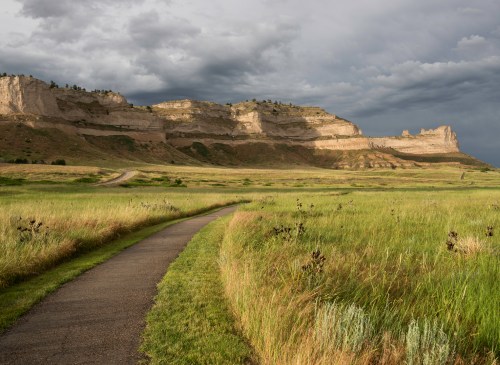 A winding path leads through tall grass towards dramatic cliffs under a cloudy sky.