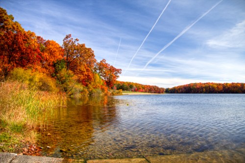 A serene lake surrounded by vibrant autumn foliage and clear blue skies, with wispy clouds overhead.