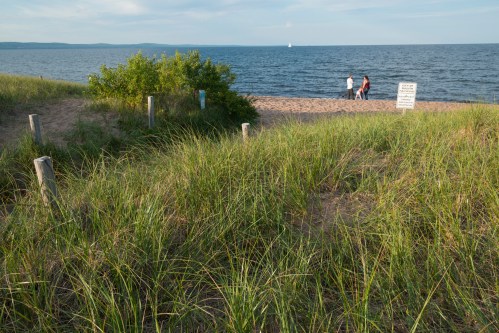 A sandy beach with tall grass in the foreground, and three people walking near the water in the background.