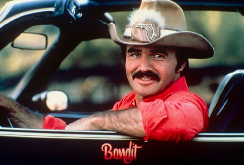 A man with a mustache and cowboy hat smiles from the driver's seat of a car, wearing a red shirt.