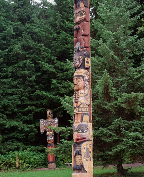 Two intricately carved totem poles stand among lush green trees in a forested area.