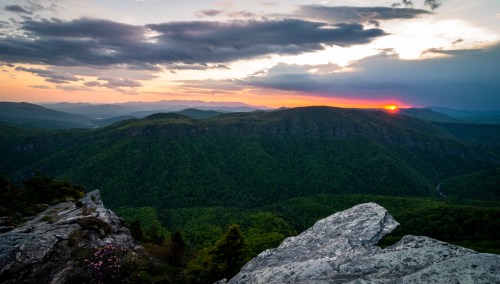 A stunning sunset over lush green mountains, with rocky foreground and dramatic clouds in the sky.