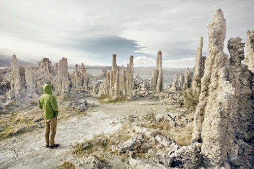 A person in a green hoodie stands among tall, rocky formations in a barren landscape under a cloudy sky.