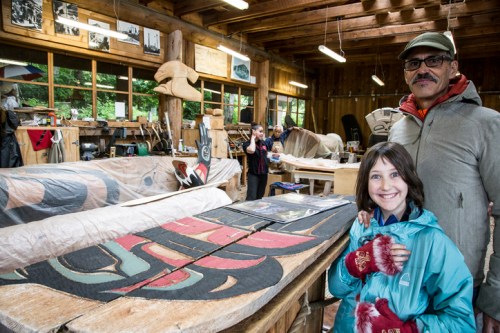 A man and a girl smile in a workshop filled with wooden carvings and tools, with people working in the background.