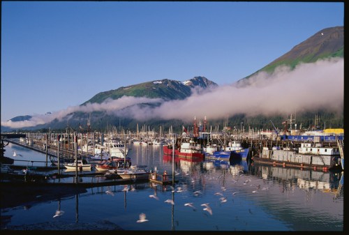 A serene harbor scene with boats docked, mountains in the background, and mist rising over the water.