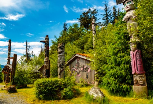 A scenic view of totem poles and a traditional wooden structure surrounded by lush greenery and blue skies.