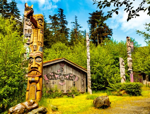 A scenic view of totem poles beside a wooden structure, surrounded by lush greenery and tall trees under a blue sky.