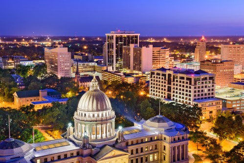 Aerial view of a city skyline at dusk, featuring illuminated buildings and a prominent dome in the foreground.